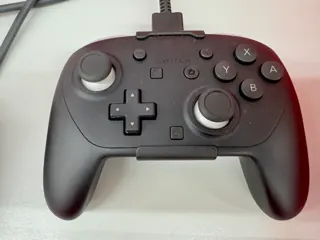 Manette Nintendo Switch 2 Pro noire et grise avec bouton C, photographiée avec un câble ; le cliché ne garantit pas le contenu du coffret