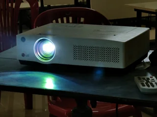 Ancien vidéoprojecteur LG blanc allumé sur une table avec une télécommande ; photo illustrative, pas la photo exacte de XGIMI MoGo 4 Laser (coffret standard) ni preuve du coffret fourni
