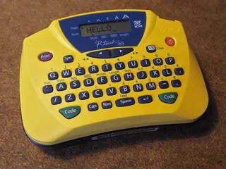 Ancienne étiqueteuse jaune Brother P-touch 65 à clavier QWERTY ; photo illustrative, pas le modèle exact Brother PT-D410 AZERTY ni le contenu de son coffret